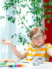 Kid boy sitting at desk clueless and confused expression with arms and hands raised. doubt concept. I don't know. Mistake. Online school lesson.
